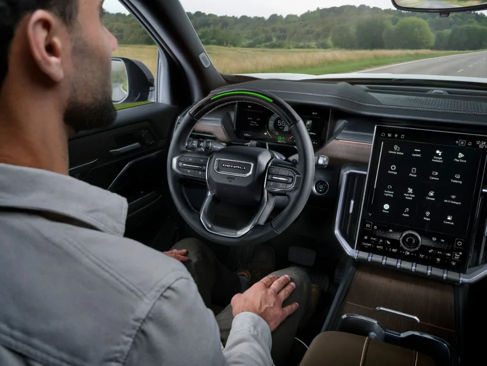 Over-the-shoulder interior view of a GMC pickup truck with a Denali-badged steering wheel, showing Super Cruise hands-free driving engaged (green steering wheel light) at 55 mph on a two-lane road