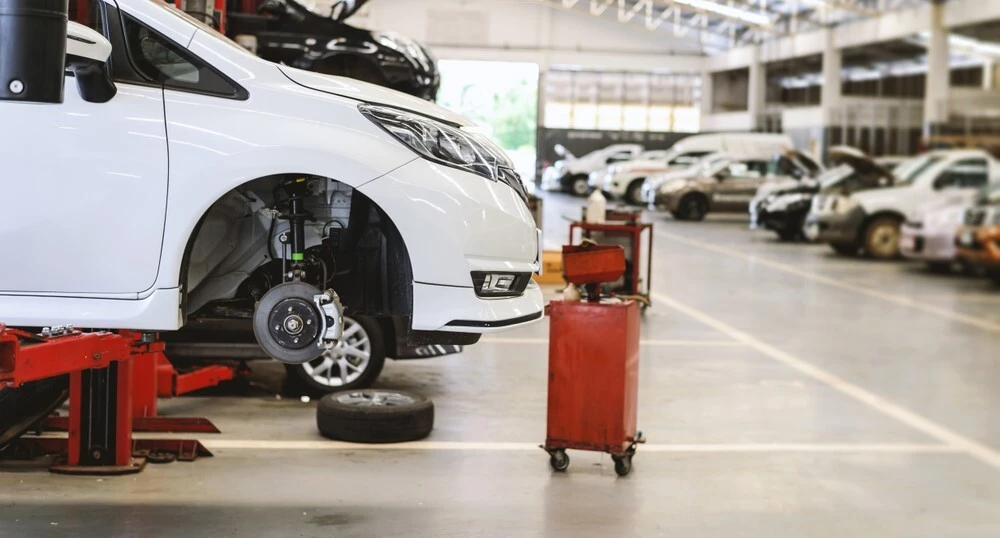 A white car with its wheels taken off is lifted onto the car jack, a mechanics tool cart sits in front of the car, off focus in the background are the other cars that are also being worked on