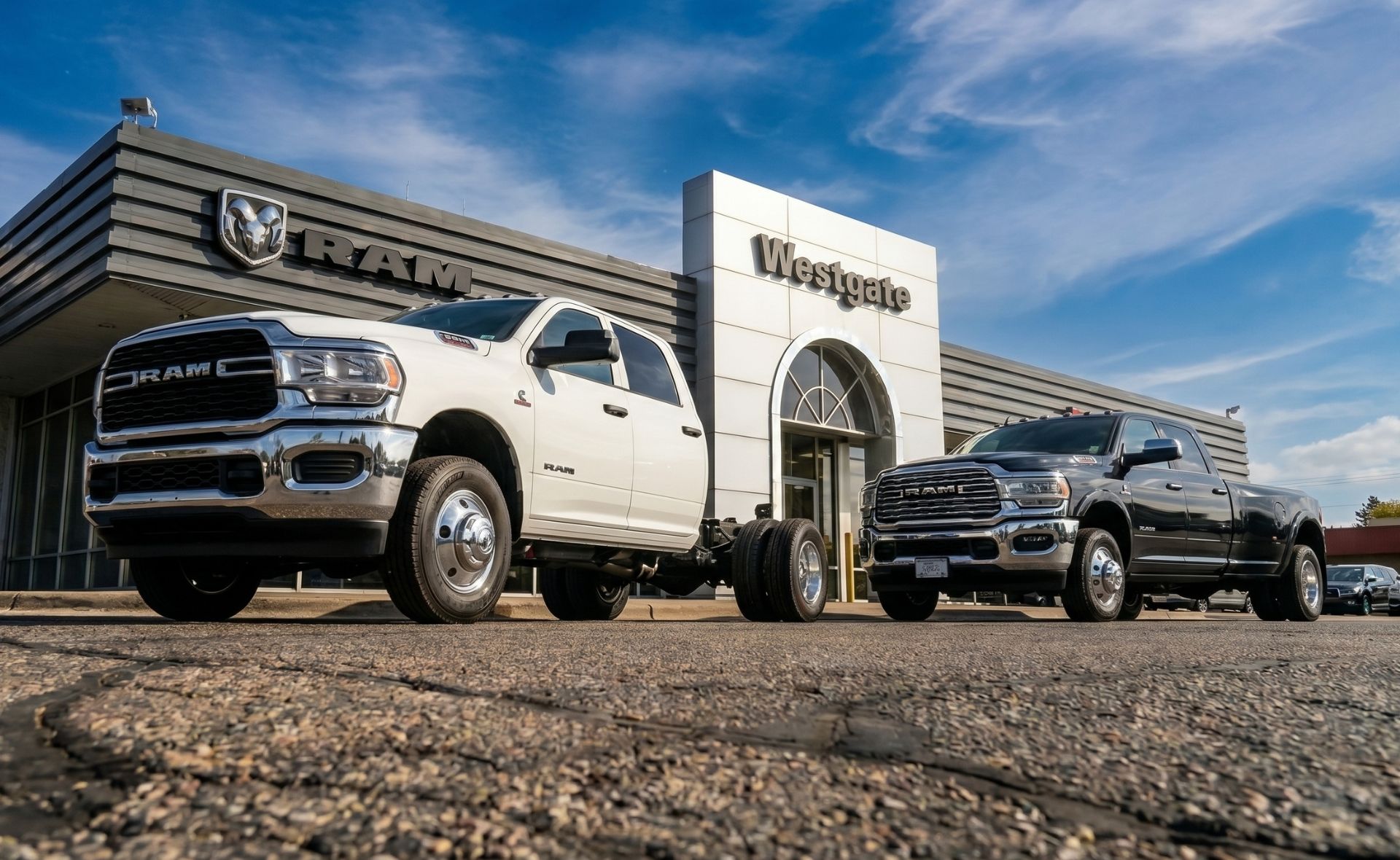 A white Ram chassis cab truck and a black Ram dually pickup truck parked side-by-side in front of the Westgate Ram dealership building in Raleigh under a blue sky.