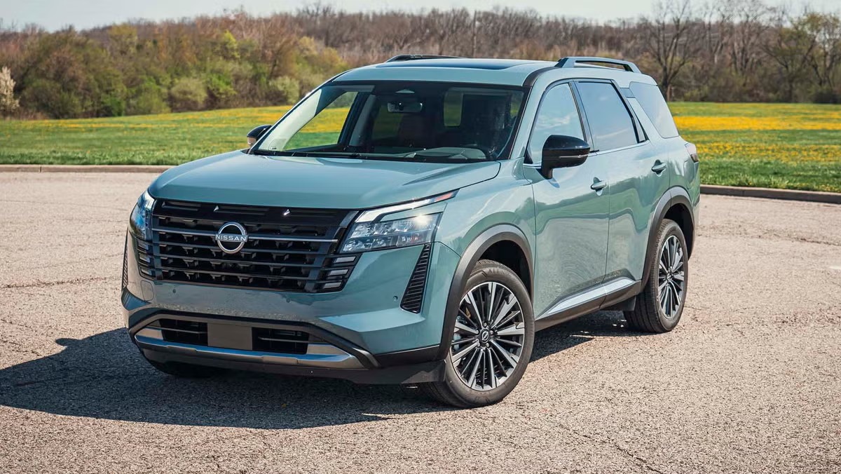 Sage green Nissan Pathfinder SUV with chrome grille and alloy wheels parked on a paved lot with a grassy field and trees in the background, shown from a front three-quarter angle.