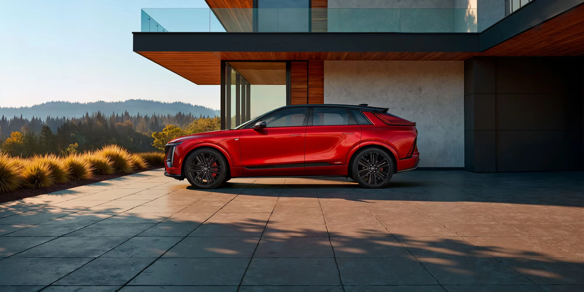 Red 2027 Cadillac Lyriq-V in side profile with black wheels and red brake calipers on a tiled driveway in front of a modern house, with forested hills and golden grass behind