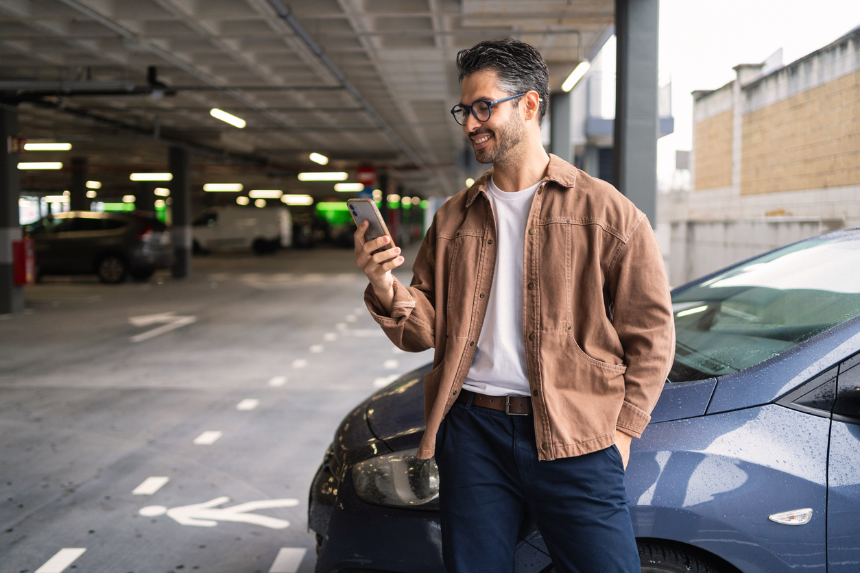 Man in casual clothes stands by a parked car in a parking garage, looking at his smartphone and smiling.