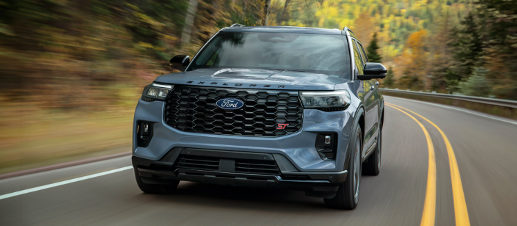 Front view of a blue-gray Ford Explorer ST driving along a winding mountain road surrounded by autumn foliage.