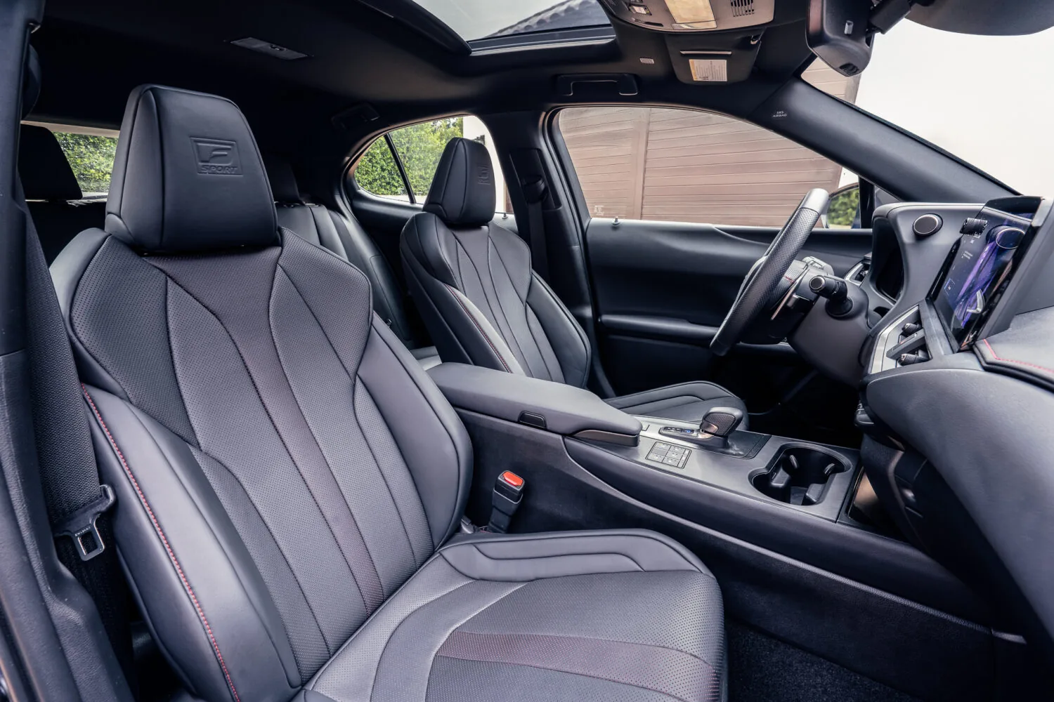 Interior view of a Lexus F Sport model showing black leather front seats with an embossed F Sport headrest logo, an open panoramic sunroof, and the center console with cupholders and gear shifter visible.