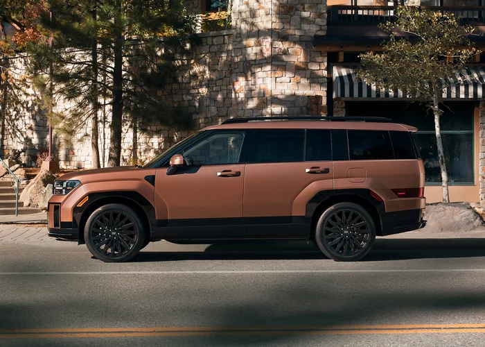 Copper 2027 Hyundai Santa Fe SUV with black wheels, black lower body cladding and roof rails, in side profile on a street in front of a stone building and autumn trees