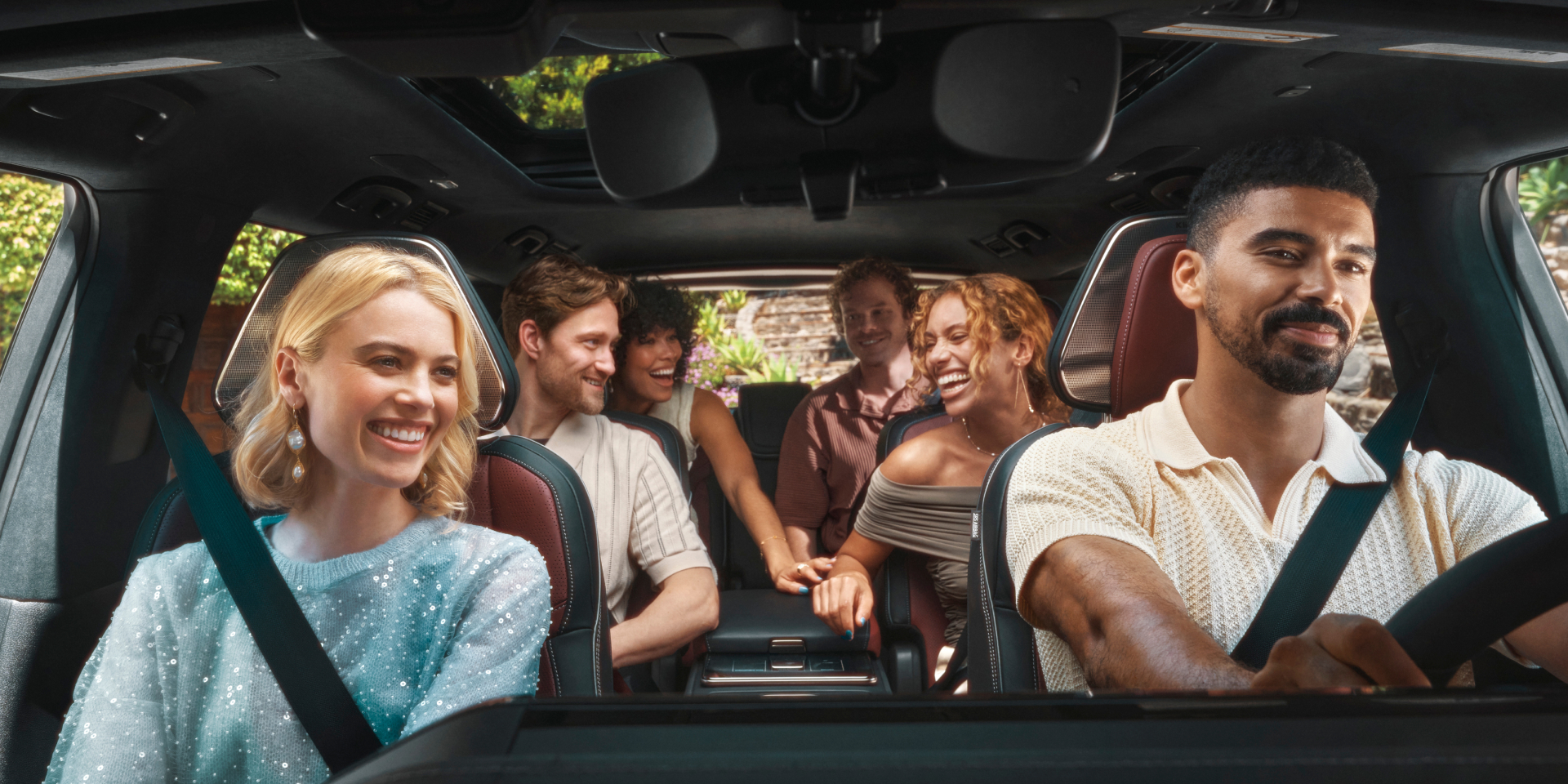 Six friends laughing together inside the spacious three-row interior of the 2027 INFINITI QX80 beneath a panoramic sunroof.