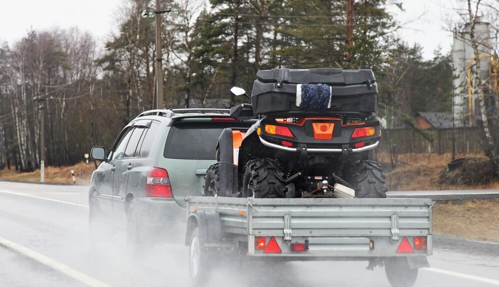 A Light Green car is towing a trailer in the foreground, on that trailer is a black and orange ATV, in the background a lot of trees are visible, the road appears as wet due to the amount of water the tires are spraying around