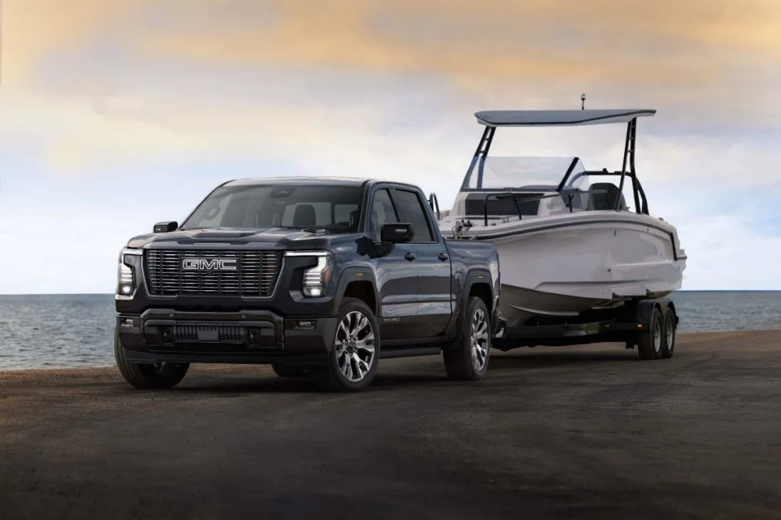A dark gray GMC pickup truck with a legible Denali badge towing a boat on a trailer along a beach at dusk, calm water in the background