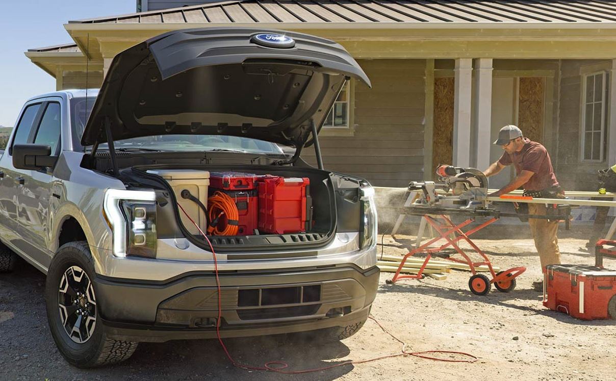 silver 2025 Ford Electric Truck with the front storage compartment open, showcasing the amount of storage space available, while a construction worker uses a saw in the background at a home construction site