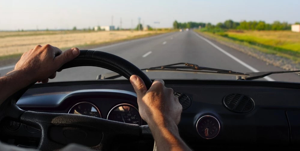 Hands of a person are visible, they are at the 10 and 2 positions on a steering wheel, the car is driving down a country road with a farm in the distance, the cars interior is black