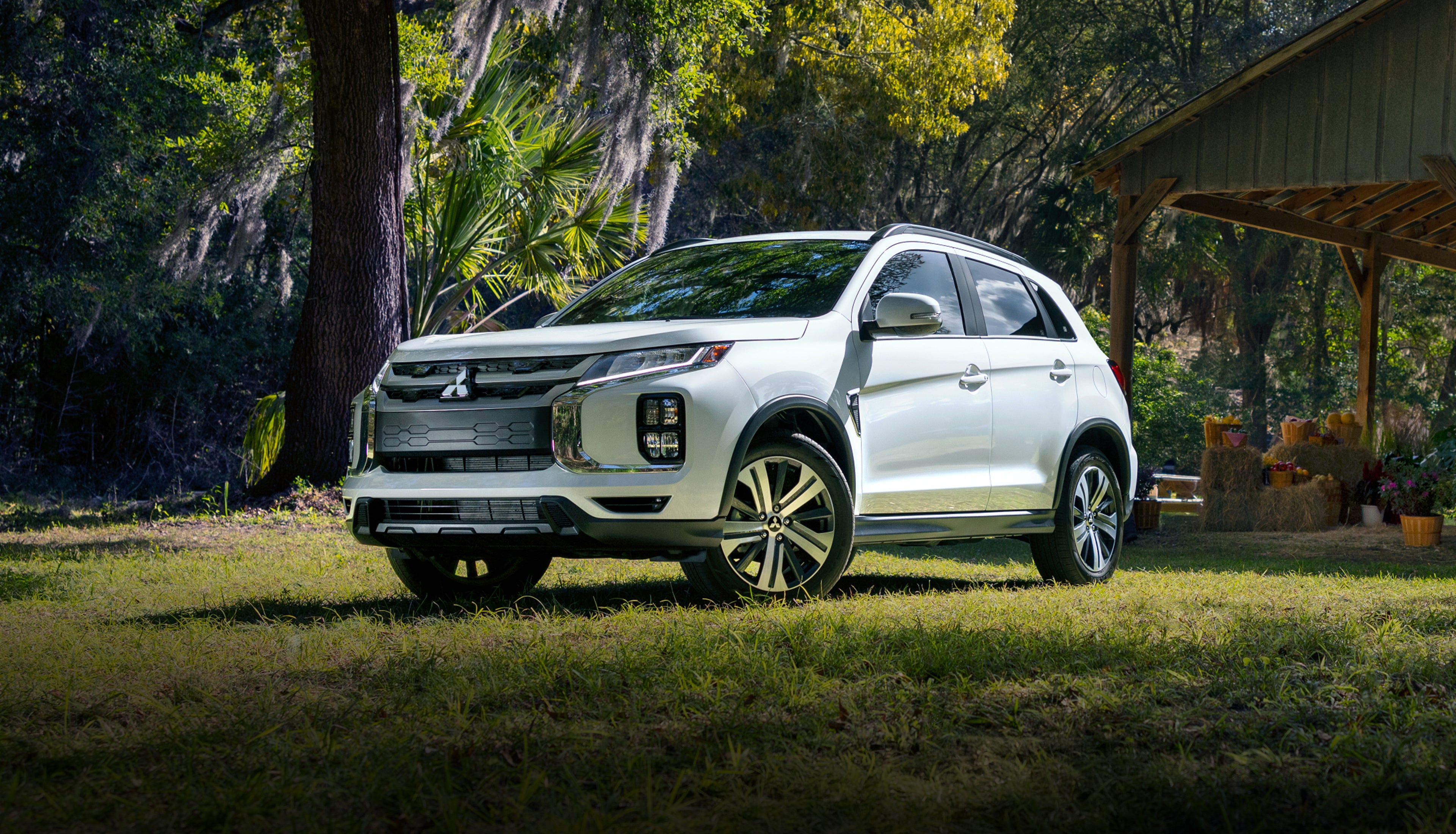 White Mitsubishi Outlander Sport SUV parked on grass beside a rustic wooden barn and farm stand, framed by trees draped in Spanish moss, shown from a front three-quarter angle.