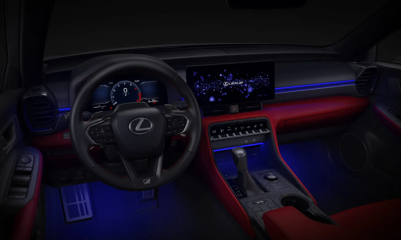  Dark Lexus cabin lit with blue ambient lighting, showing a black steering wheel with the Lexus emblem and an F SPORT badge on the lower spoke, a digital gauge cluster, a wide touchscreen showing the Lexus logo, red leather trim, and a floor-mounted shifter.