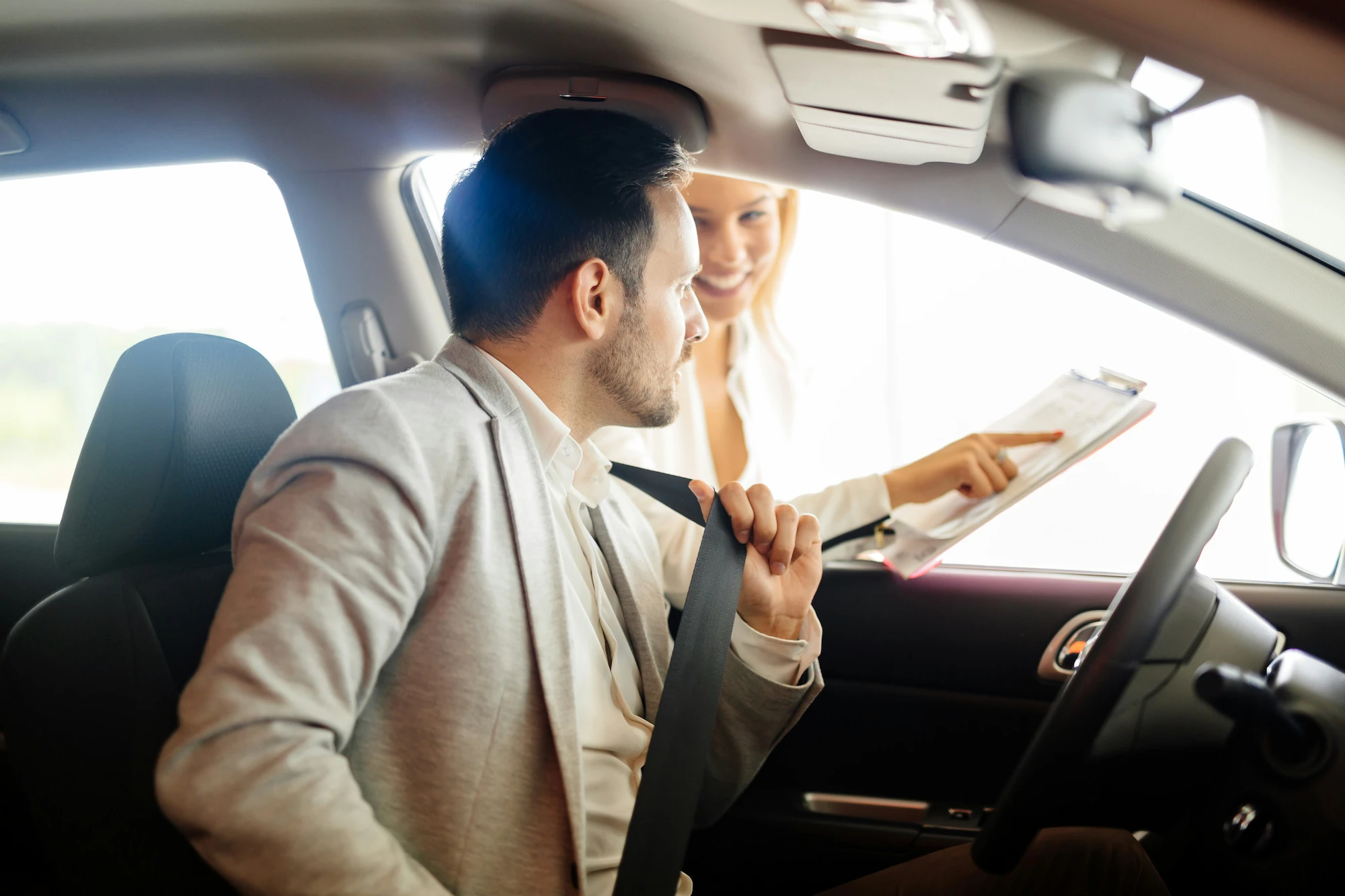Salesperson pointing out details on a clipboard to a customer buckling his seatbelt before a test drive