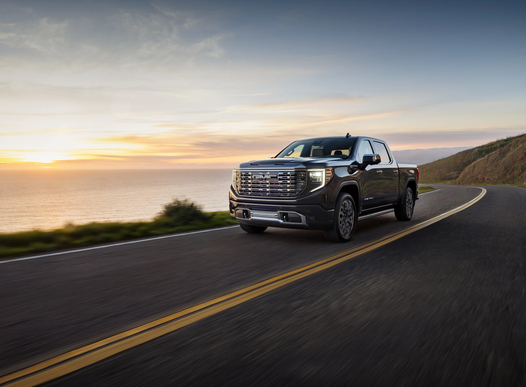 A black GMC Denali pickup truck drives along a coastal road at sunset, with the ocean and cliffs in the background