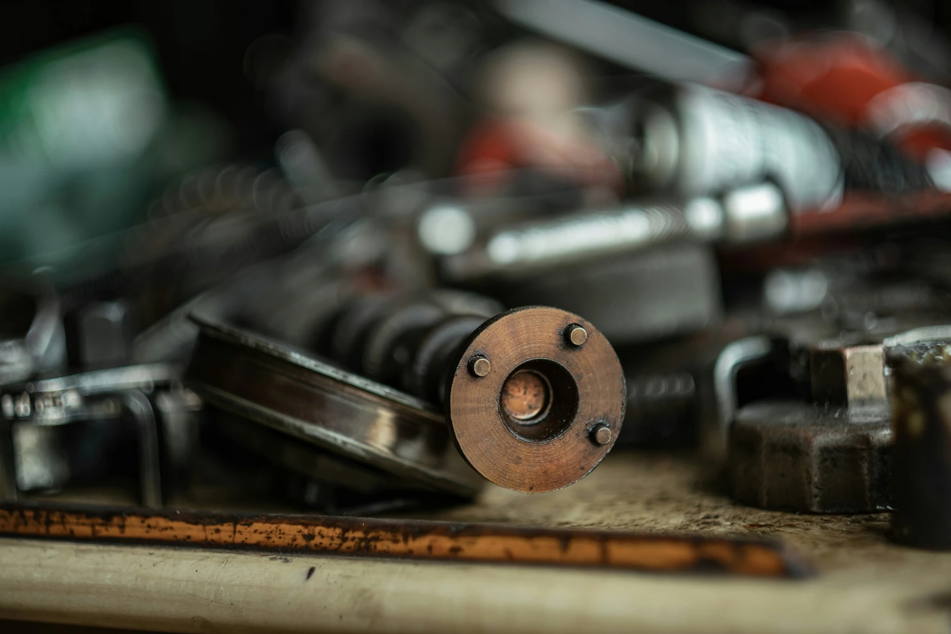Macro close-up of a copper-toned cylindrical mechanical component with three bolt heads, resting on a wooden ruler on a workbench, with blurred tools and parts in the background