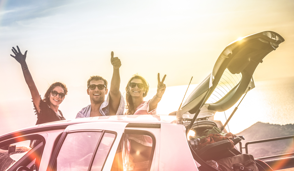 White car with its door open and trunk open with road tripping equipment packed into the trunk, there are 3 people hanging around the car, one is waving, one is giving a thumbs up , and the other is giving a peace sign. There is sun in the background