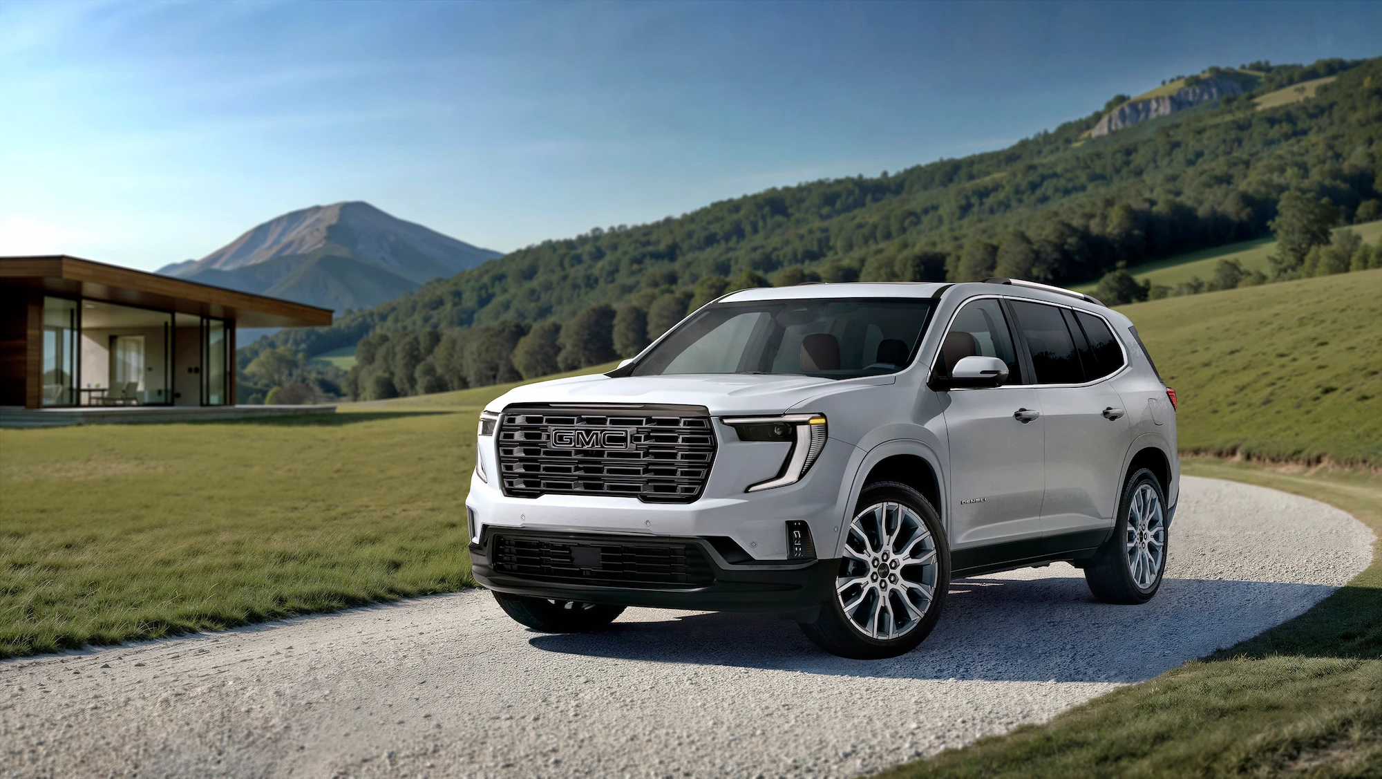 Front three-quarter exterior view of a 2026 GMC Acadia Denali Ultimate in Glacier White Tricoat, parked on a gravel driveway in front of a modern house with mountains in the background