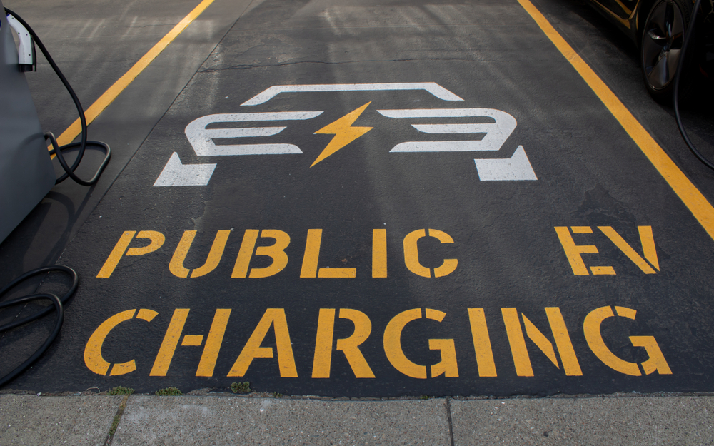 Parking spot with the words Public EV Charging in orange along the bottom of the spot, a graphic of the back of a white car with an orange  electric bolt through the middle is above the words below it, an ev charing cord is visible on the far left of the image