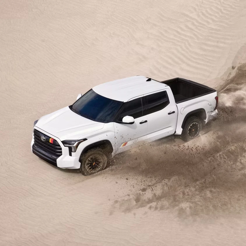 White Toyota Tundra crew cab pickup with black wheels and retro-style stripe decals, seen from above driving through a sand dune and kicking up a plume of sand