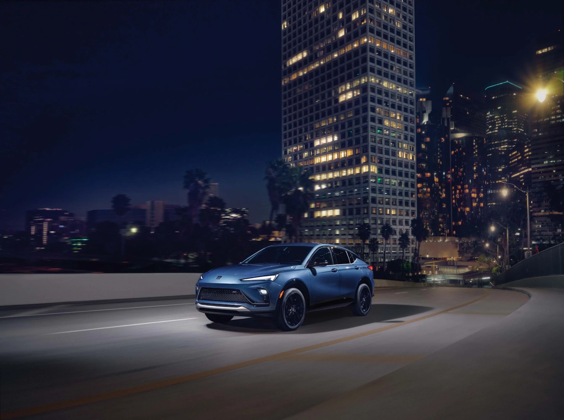 A blue SUV drives along an elevated city roadway at night, with illuminated skyscrapers in the background