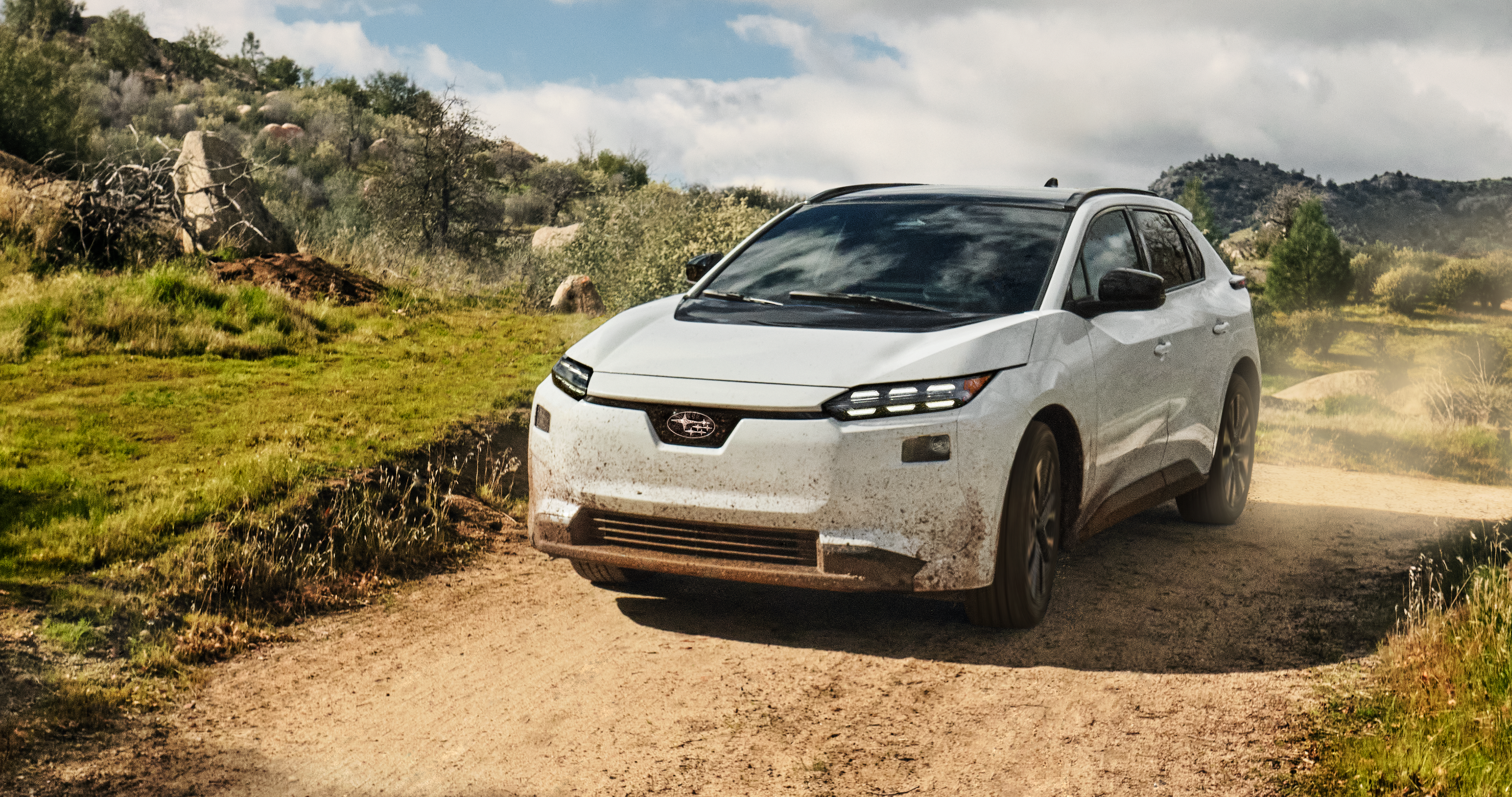26_SOL_overview_hero_md_sm.png & 26_SOL_overview_hero_xl_lg.png / A white 2026 Subaru Solterra electric SUV drives on a dirt road in arid hills, kicking up dust behind.