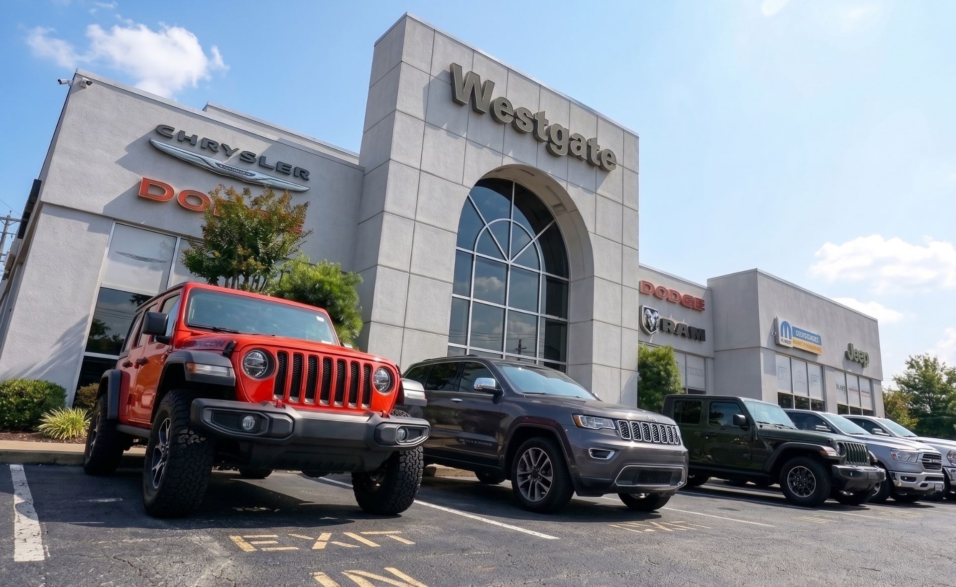 An orange Jeep Wrangler Unlimited, a grey Jeep Grand Cherokee, and a dark green Jeep Wrangler parked in front of the Westgate Chrysler Jeep Dodge Ram dealership building entrance on a clear, sunny day in Raleigh.