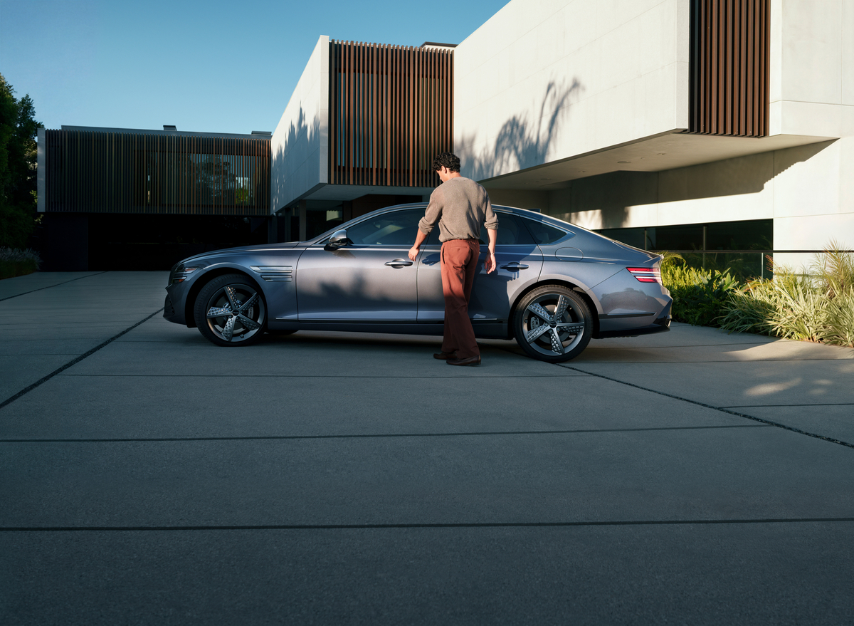 Driver side profile view of driver reaching for door handle on Genesis G80 parked beside modern home