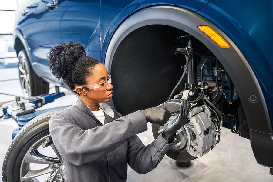 Female technician wearing safety glasses works on a brake caliper and wheel hub on a blue SUV with the wheel removed, in a service bay WCAG AA Notes: Distinguished from the existing brake rotor/caliper close-up
