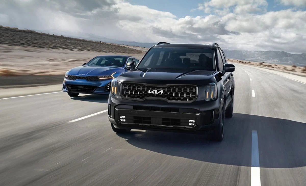 Black Kia SUV and blue Kia sedan driving side by side on a desert highway under a cloudy sky