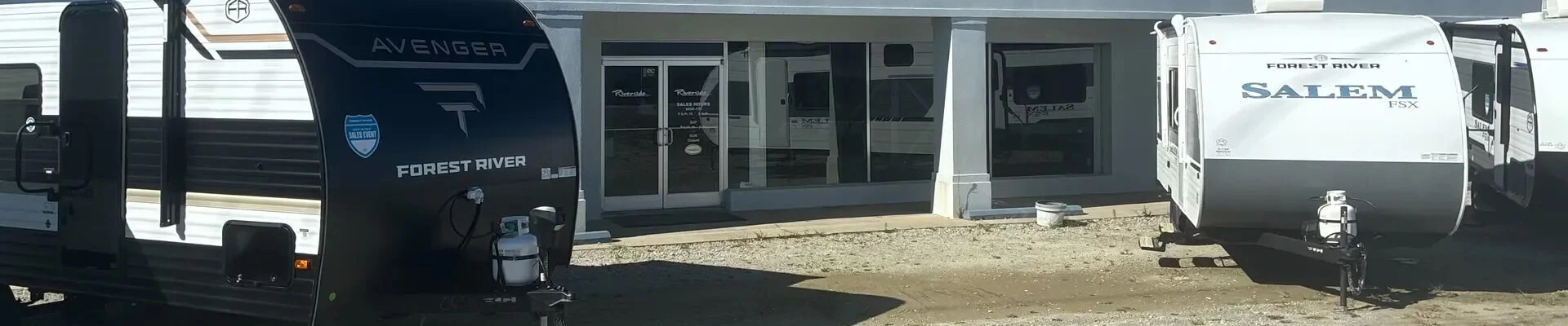 Exterior view of black Forest River Avenger and white travel trailers outside Riverside RV dealership.