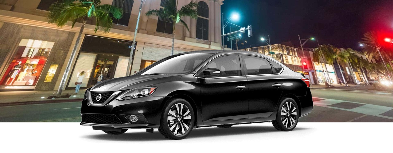 Black 2017 Nissan Sentra sedan in front of a palm-lined shopping street at night