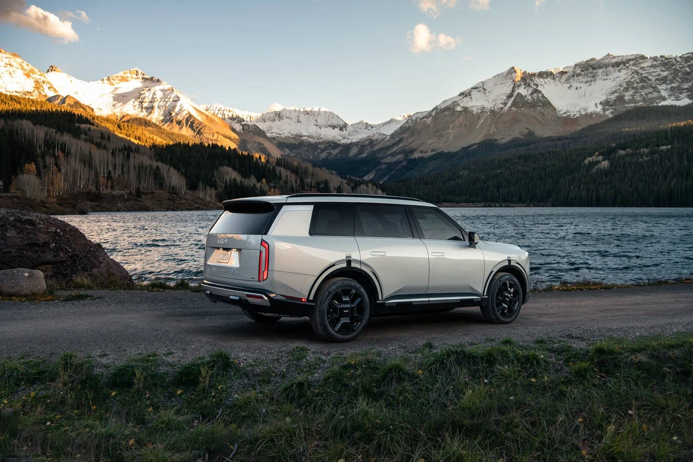 Silver 2027 Kia Telluride SXP, rear three-quarter view, parked beside a lake with snow-capped mountains and pine forest behind it.