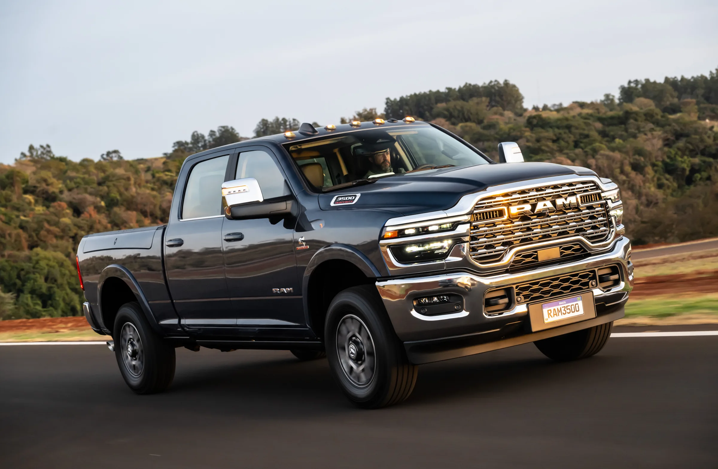 2026 Dark blue-gray Ram 3500 Heavy Duty pickup truck driving on a paved road with tree-covered hills in the background