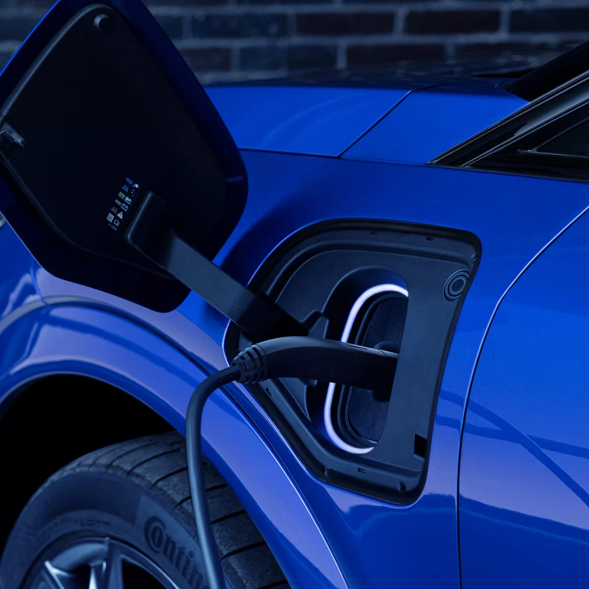 Close-up of the open charge port on a blue Cadillac Optiq-V with a charging connector plugged in and a white light ring glowing around the port, next to the front tire
