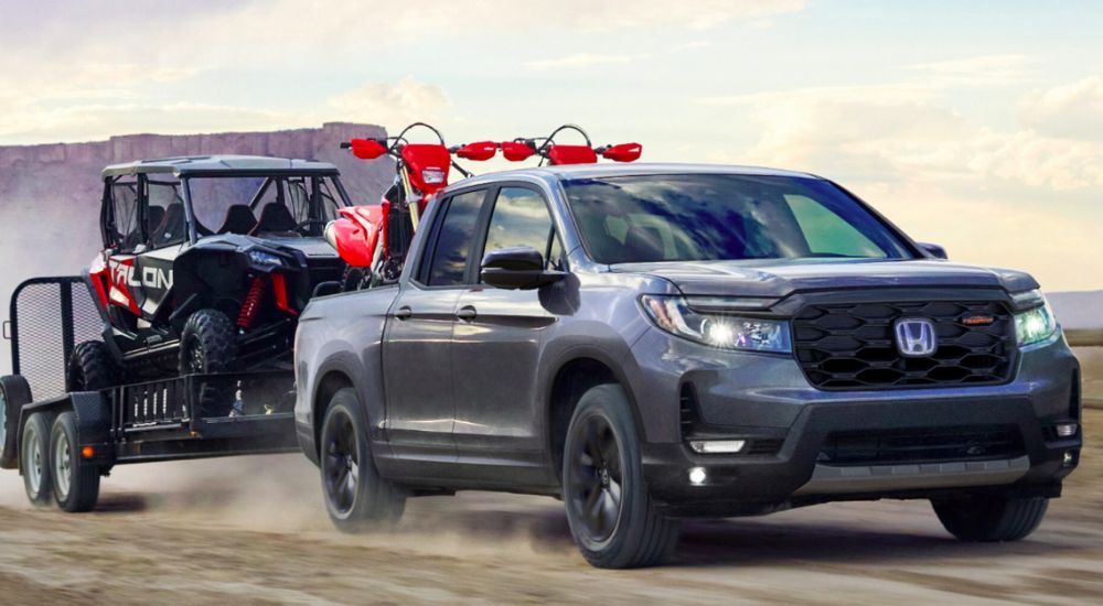 Grey 2026 Honda Ridgeline TrailSport driving off-road towing ATVs.