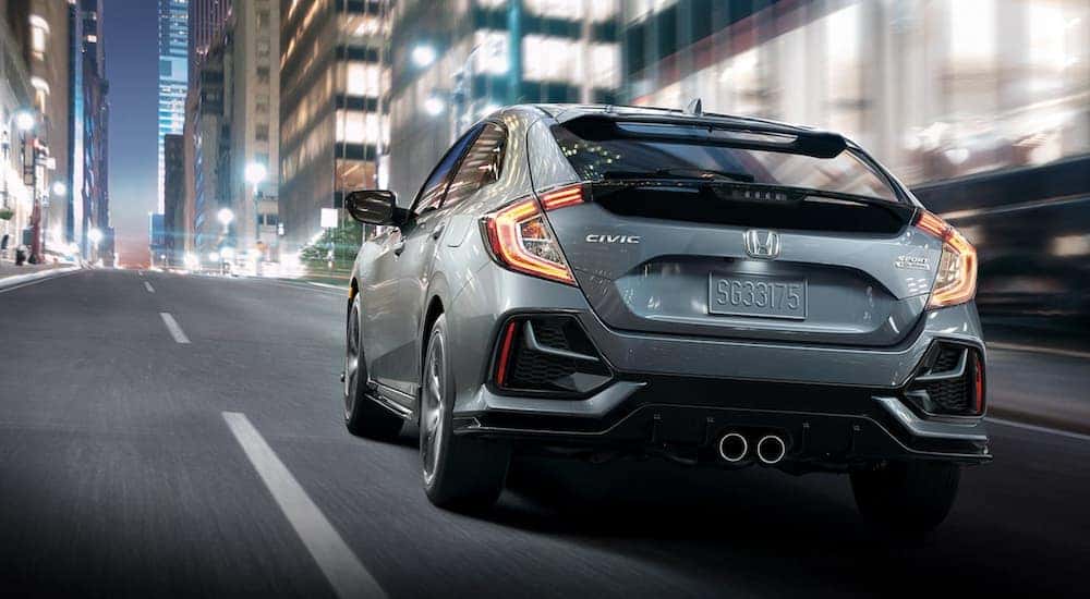 A grey 2020 Honda Civic Hatchback is shown from a rear angle while it drives down a city street.