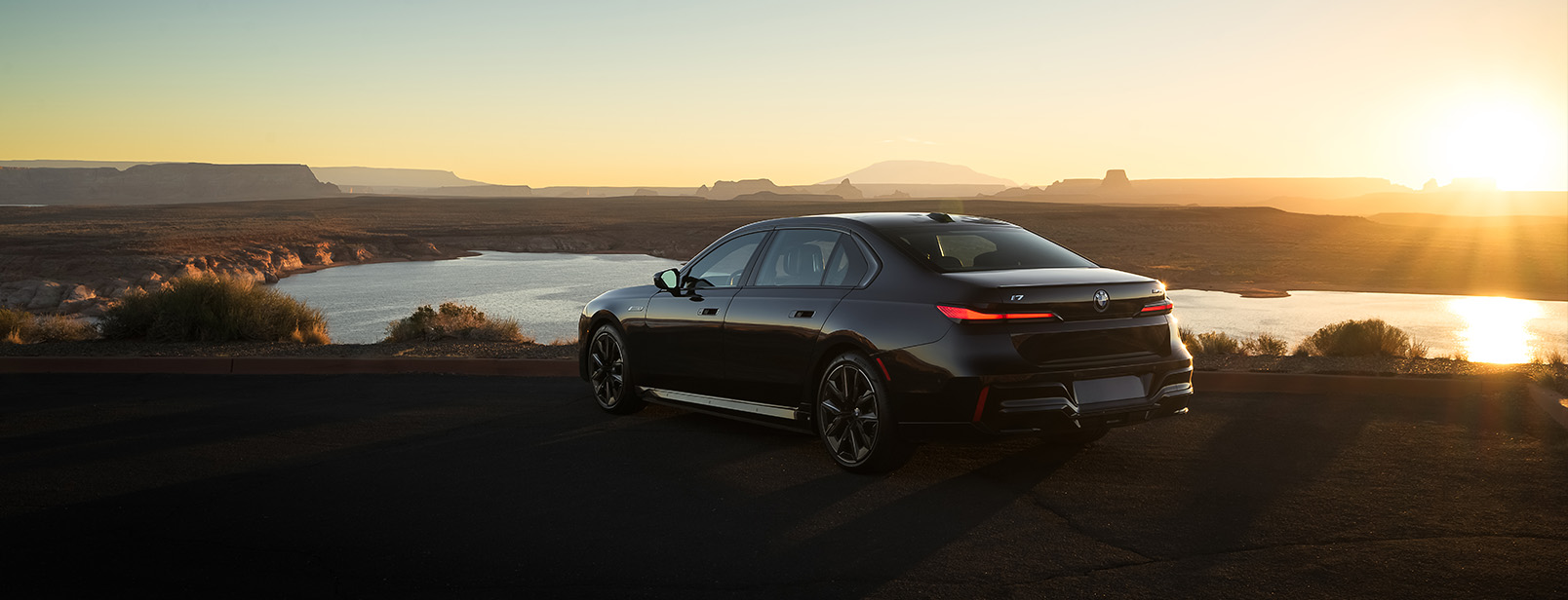 Rear and side profile of a black 2026 BMW i7 M70 parked in flat, desert landscape with the sun setting in the background. 
