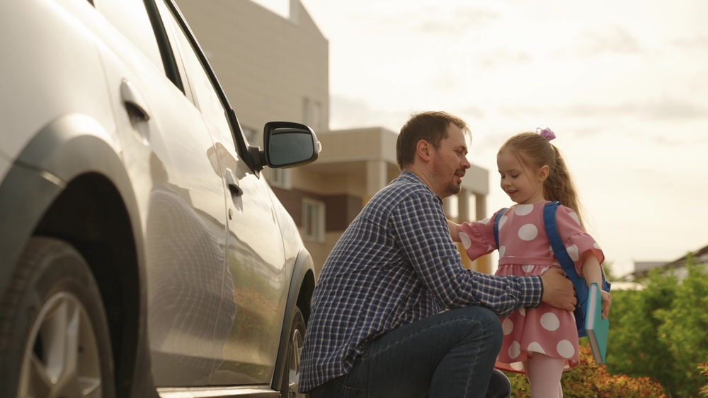 Father kneeling and smiling while talking to his daughter, who is wearing a pink polka dot dress and a blue backpack, standing next to a car.