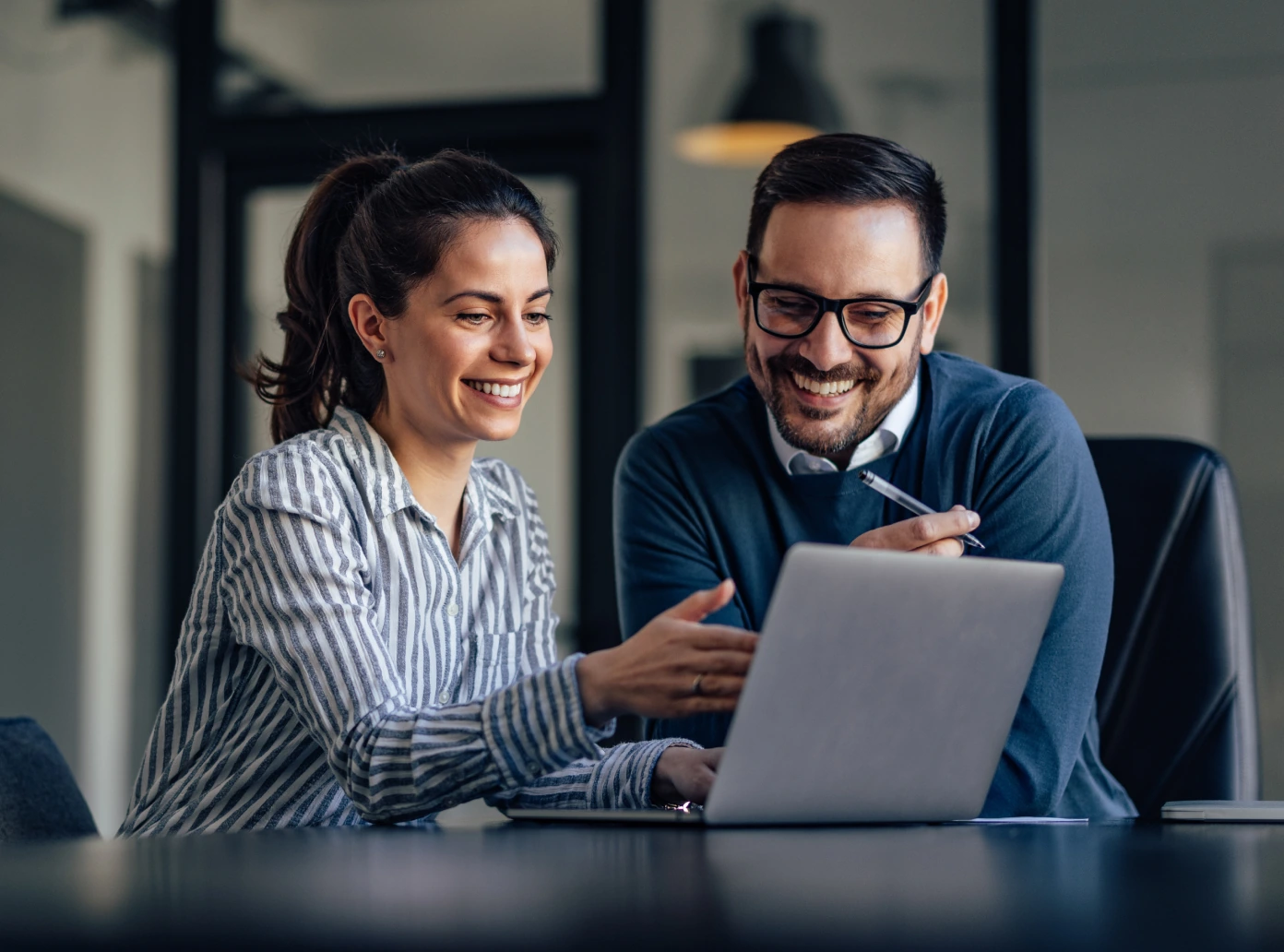 A smiling man and a woman sitting at a table with a laptop. The woman points to the screen.