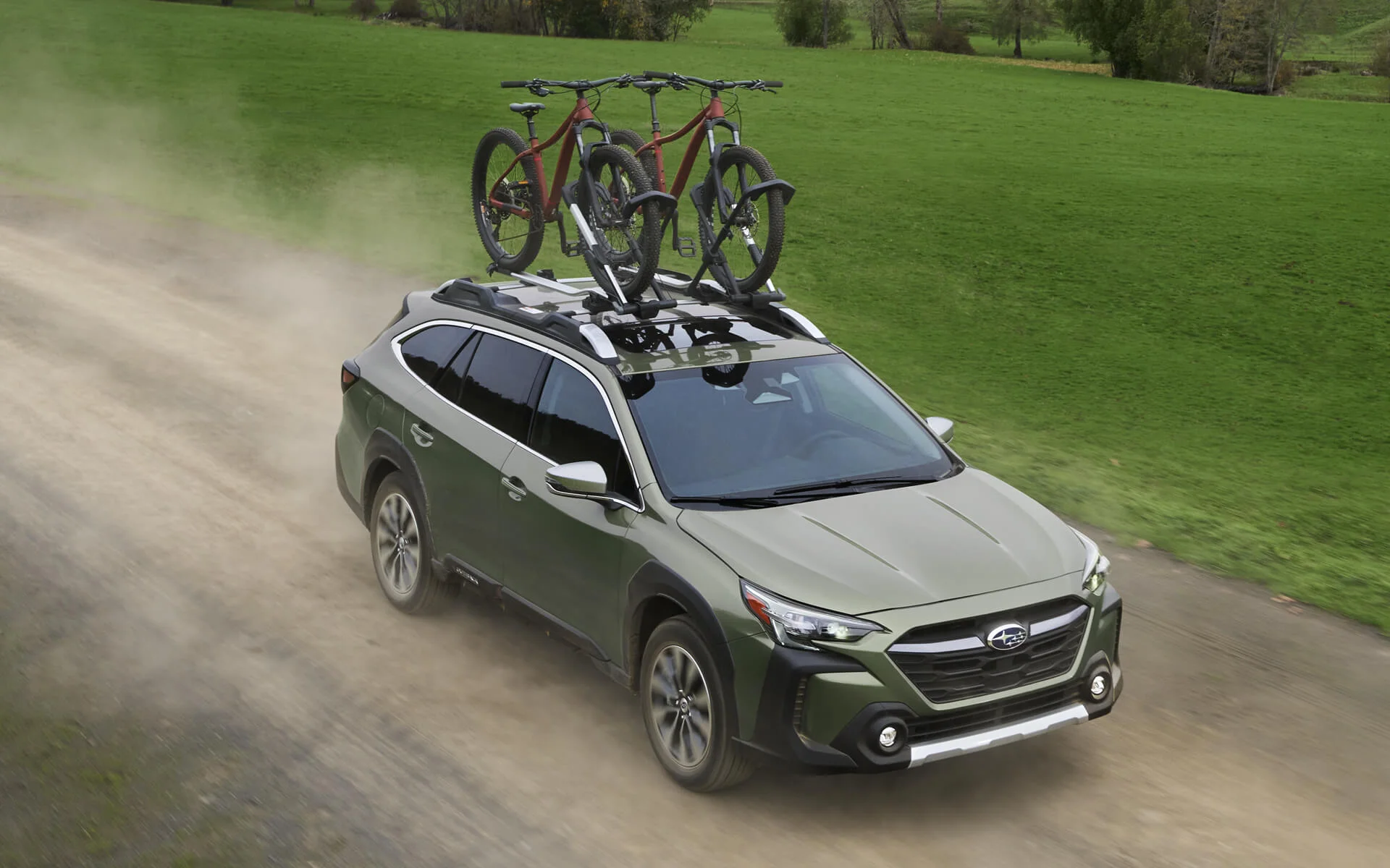 Green 2023 Subaru Outback with bikes on a roof rack driving on a dirt road