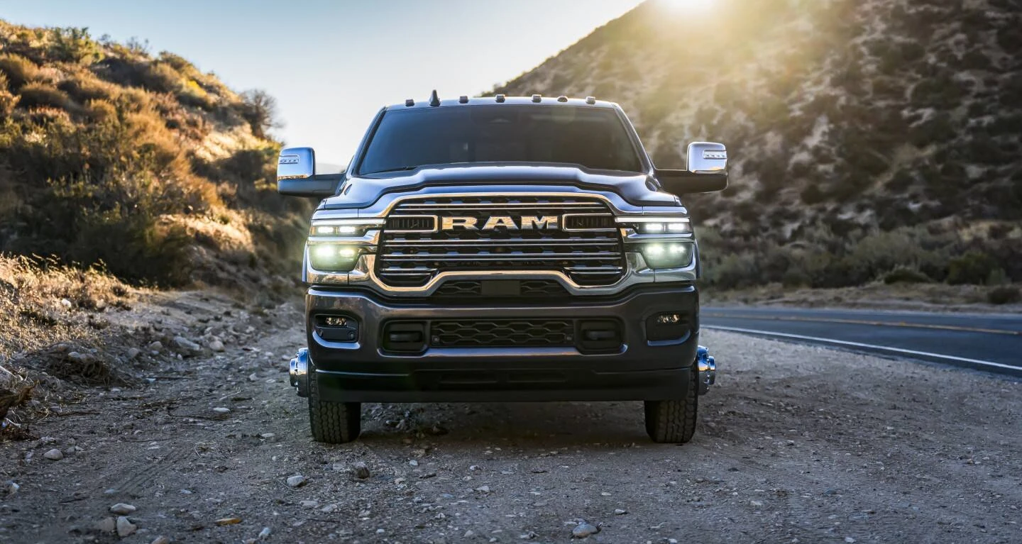 Dark gray 2027 Ram 3500 heavy-duty pickup with a chrome grille, RAM lettering, large tow mirrors, roof marker lights and headlights on, facing the camera on a gravel pullout beside a mountain road with the sun glaring over a hillside