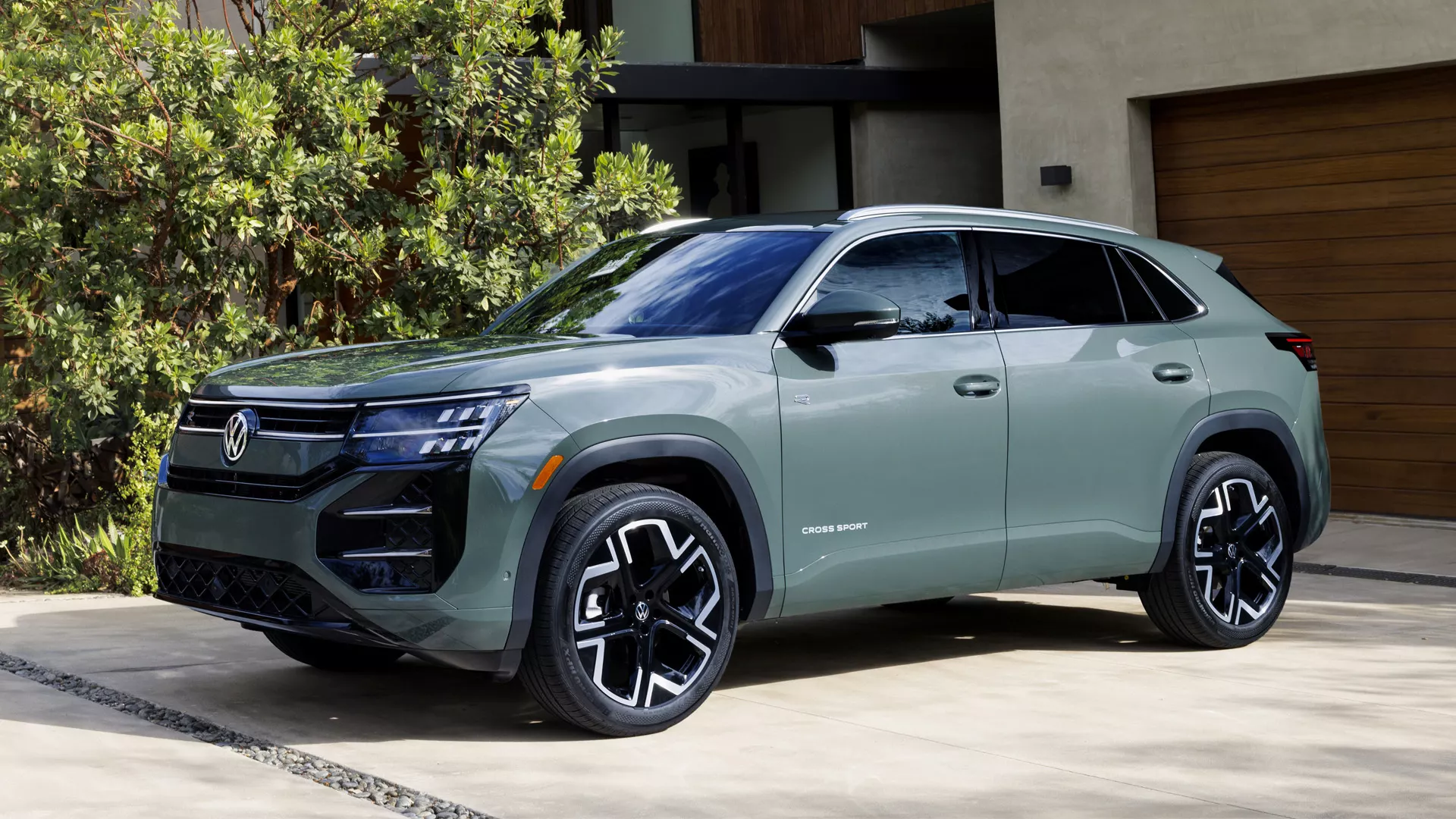 Green Volkswagen Atlas Cross Sport SUV with black alloy wheels and R-Line styling parked in a paved driveway in front of a modern home, shown from a front three-quarter angle.