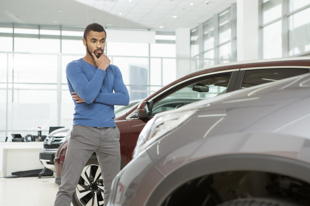 A man looks onto a silver car of which we can only see the hood, behind him are two other cars and behind those 2 cars a desk, the setting appears to be a car dealership