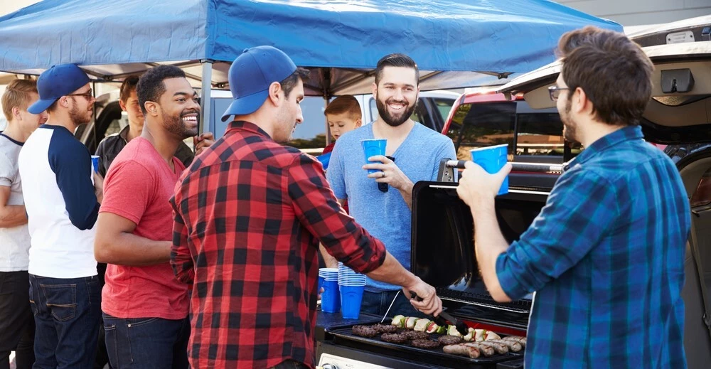 A Group of men are in tailgating in a parking lot, theyre holding blue solo cups and are grilling various meats, a canopy is constructed in the background