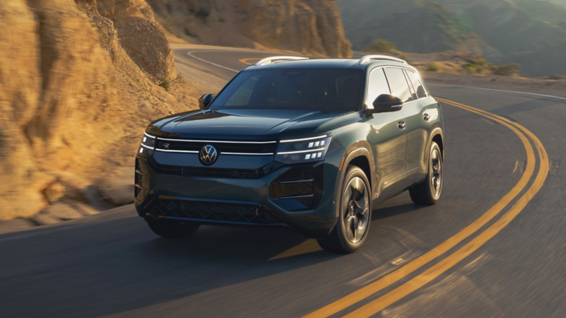 Dark green 2027 Volkswagen Atlas SUV with R-Line badging and an illuminated light bar grille driving along a curving two-lane mountain road, shown from a front three-quarter angle.
