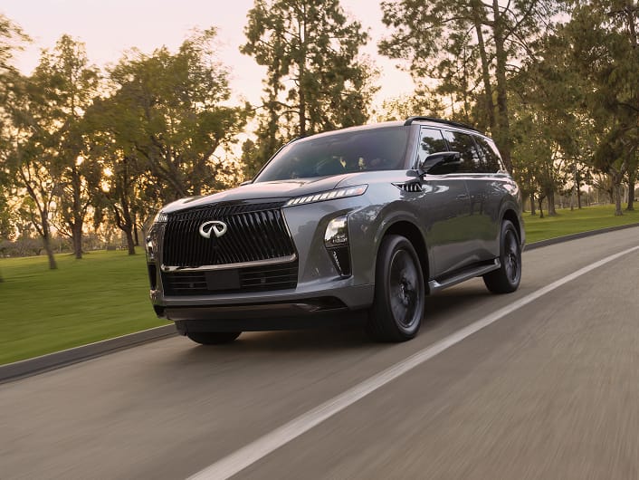 Front three-quarter view of a metallic gray 2027 INFINITI QX80 driving on a tree-lined road in golden-hour light.