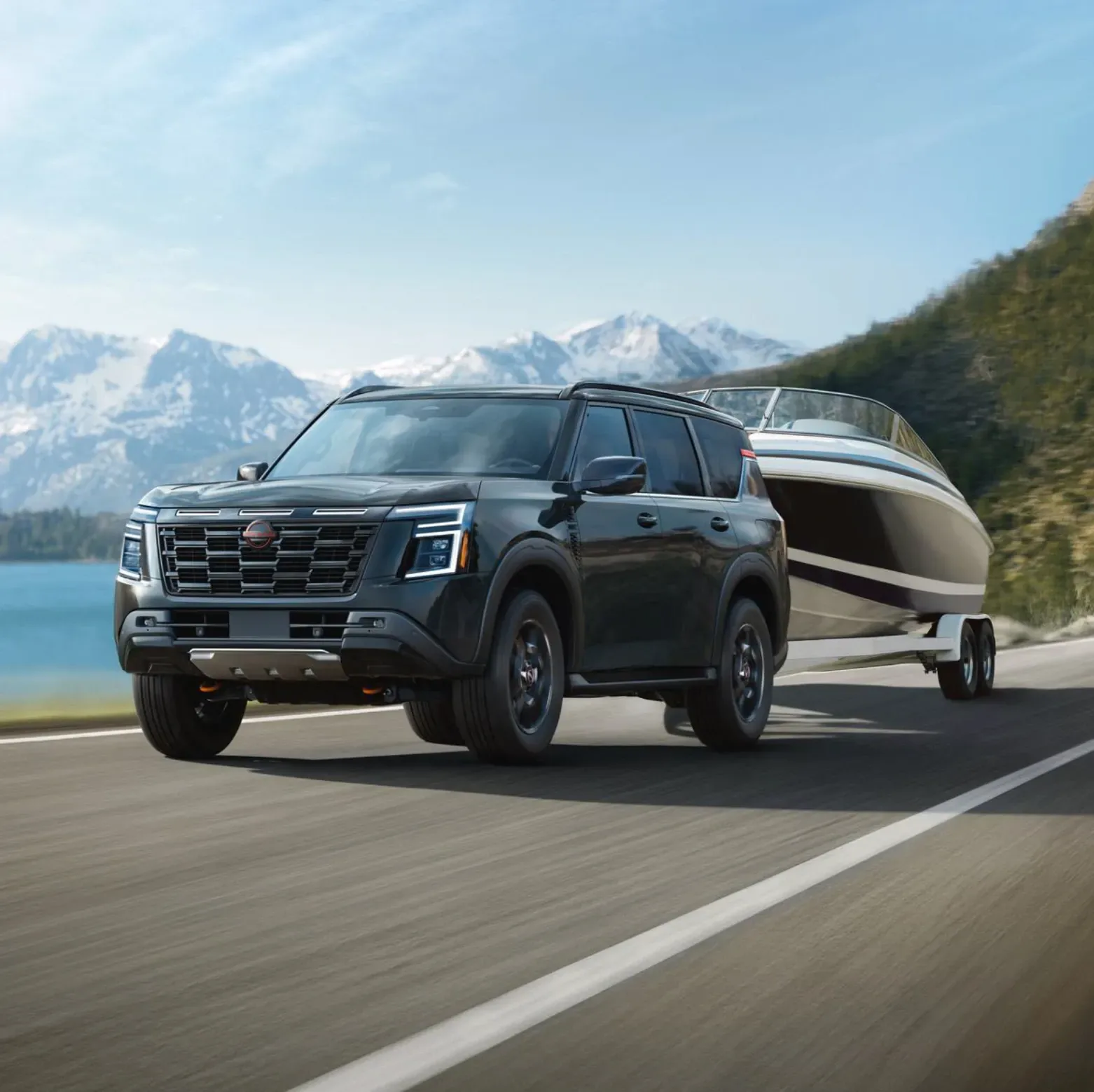 Black Nissan SUV towing a white boat on a trailer along a lakeside highway, snow-capped mountains and a forested hill behind