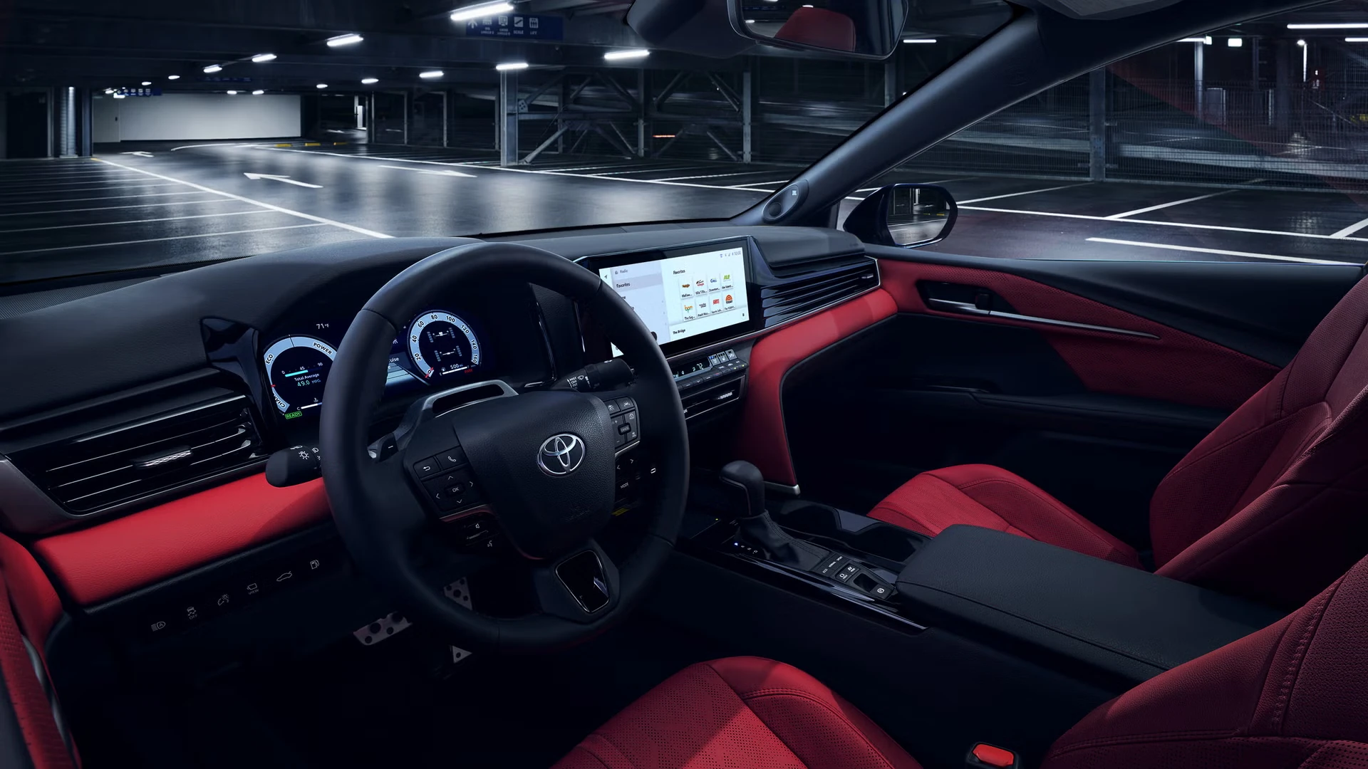 Interior of a Toyota Camry with red perforated seats, red dashboard and door trim, a black steering wheel with the Toyota emblem, a digital gauge cluster and a center touchscreen, in a parking garage