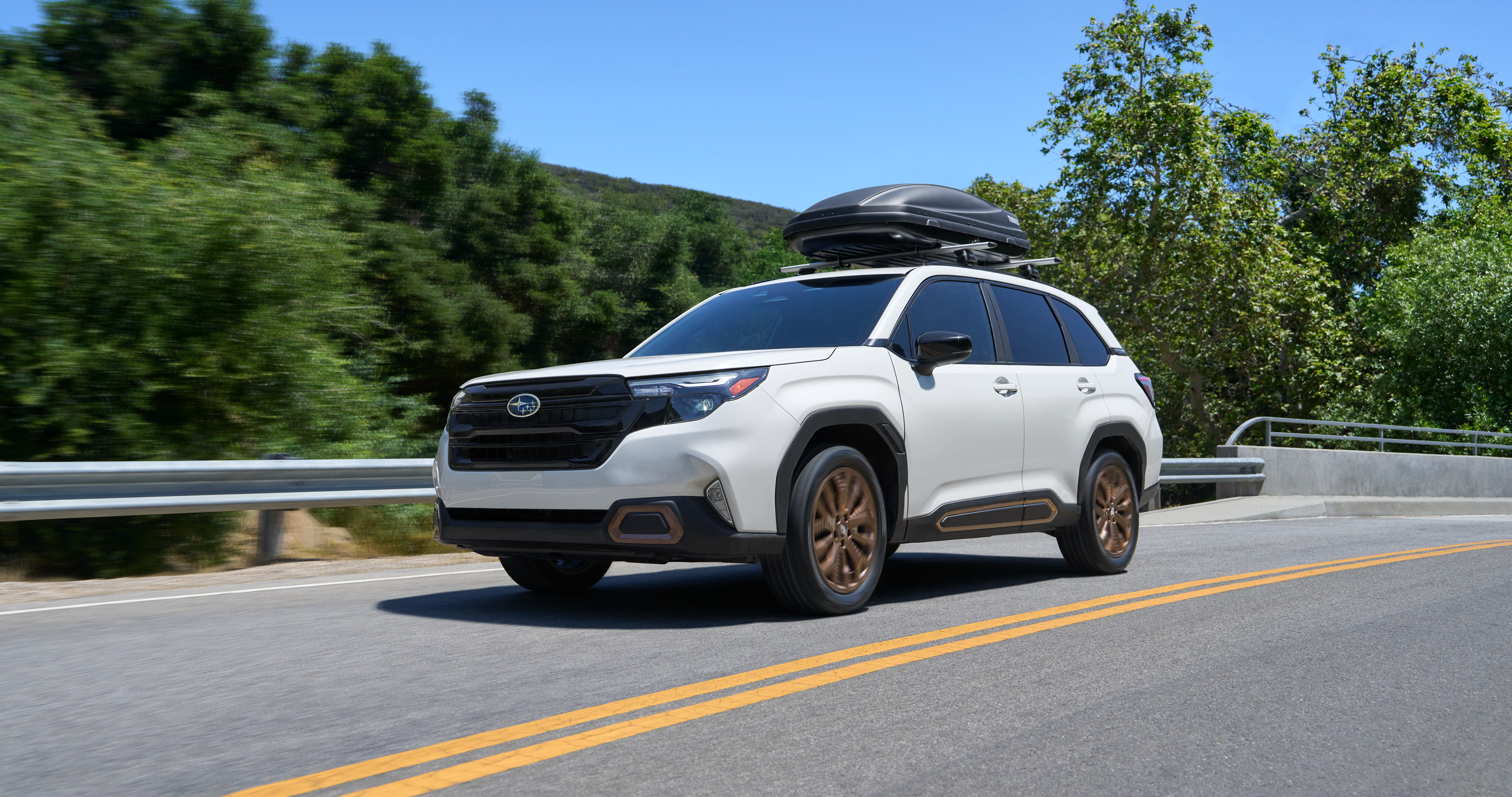 26_FORA_hero_gallery_1.png / A 2026 Subaru Forester AWD SUV with a Thule storage attachment on its roof rails driving on a highway. 
