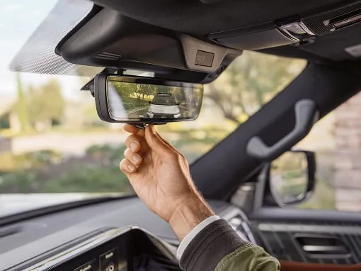 Close-up of a hand adjusting the smart rearview mirror in a 2027 INFINITI QX80; the digital display shows a clear camera feed of the road and a white SUV behind the vehicle.